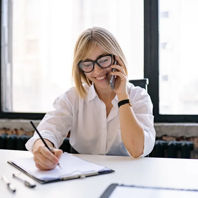 Smiling employee on the phone, taking notes at her desk